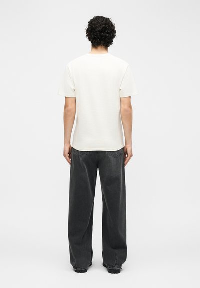 Άτομο με κατσαρά μαλλιά που φοράει απλό λευκό κοντομάνικο μπλουζάκι και φαρδιά μαύρα τζιν, στέκεται γυρισμένο με την πλάτη σε λευκό φόντο.