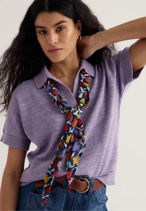 Ženska z dolgimi temnimi lasmi, oblečena v sivkasto kratka pletena majica, cvetlični šal zvezan okoli ovratnika, rjav pas in modre kavbojke, ki pozira z dvignjeno roko.