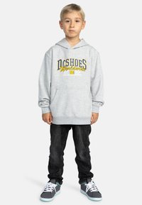 Grijze hoodie met een voorzak in kangoeroe-stijl, voorzien van de opdruk "DC Shoes Worldwide" in blauw en geel. Gepaard met zwarte jeans en grijze sneakers.