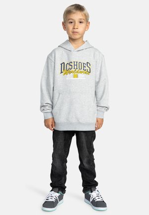Grijze hoodie met een voorzak in kangoeroe-stijl, voorzien van de opdruk "DC Shoes Worldwide" in blauw en geel. Gepaard met zwarte jeans en grijze sneakers.