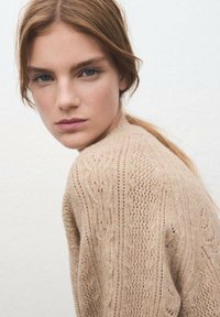 Suéter de punto en color beige claro con un patrón texturizado, que presenta detalles de calado y un corte relajado. La tela parece suave y acogedora.