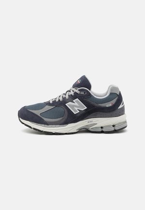 Blauer und grauer New Balance Sportschuh mit weißem "N"-Logo, gepolsterter Sohle und Schnürsenkeln auf hellgrauem Hintergrund, nach links gerichtet.