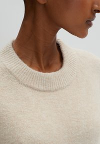 Pull en laine beige avec un col rond large et côtelé, et une surface douce et texturée. Aucun motif visible ni détail matériel.