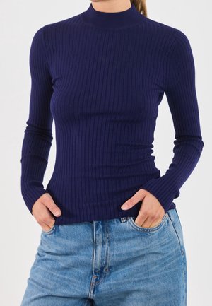 Femme portant un pull bleu marine ajusté à côtes, manches longues, et un jean taille haute bleu clair avec les mains dans les poches.