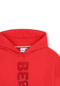 Felpa rossa con cappuccio e una grafica centrale "BE" in una tonalità più scura, realizzata in un materiale morbido con una texture liscia.