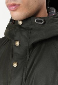 Σκούρο πράσινο μπουφάν με ψηλό γιακά, που διαθέτει χρυσά κουμπιά, κλεισίματα με κορδόνια και καρό επένδυση. Λείος υφασμάτινος ιστός, ελαφρώς ανθεκτικός στο νερό.