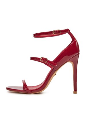 Czerwony sandał na szpilce z lakierowanej skóry z trzema cienkimi paskami i małymi złotymi klamrami na białym tle.
