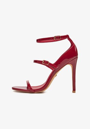 Sandalia de tacón de aguja de charol rojo con tres tiras delgadas y pequeños broches de hebilla dorada sobre fondo blanco.