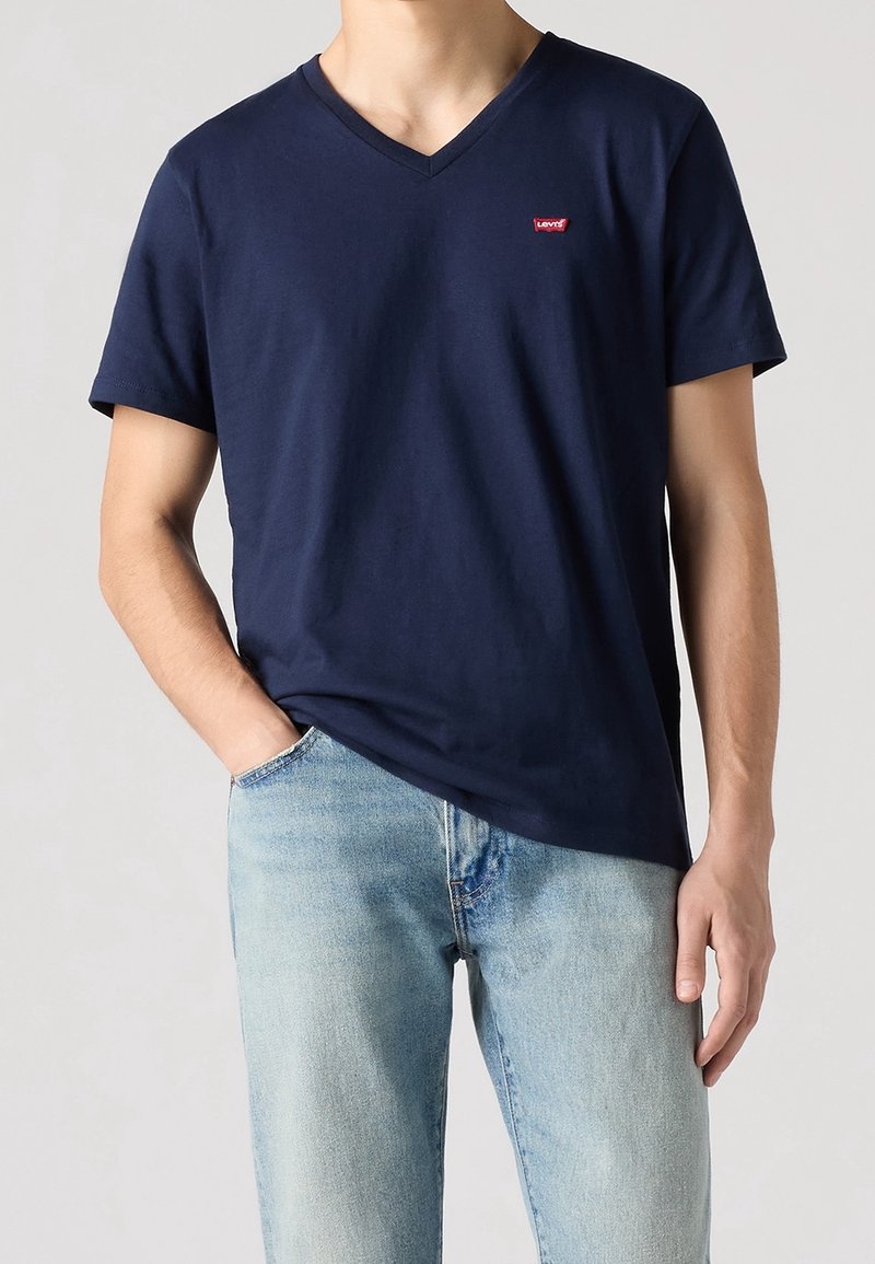 Homme portant un T-shirt Levi's bleu marine col V et un jean bleu clair, main dans la poche, sur fond gris uni.