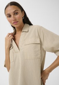 Beige randig skjorta med krage, korta ärmar och två bröstfickor. Tyget verkar ha en texturerad yta med en lätt design.