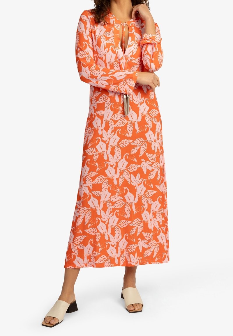 Femme portant une robe maxi orange à manches longues avec imprimé feuilles et oiseaux roses, col à nouer avec pompons et sandales ouvertes à talons blocs beige.