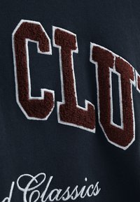 Marinblå sweatshirt med djärva broderade bokstäver i bordeaux "CLUB" med vita accenter. Mjuk textur och klassiska kollegiala designelement.