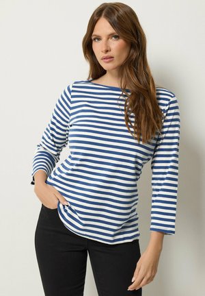 Donna con lunghi capelli castani che indossa una camicia a righe blu e bianche a maniche lunghe e pantaloni neri, in piedi con una mano in tasca.
