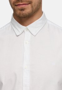 Homme portant une chemise blanche boutonnée avec un col pointu et un logo brodé subtil sur la poitrine, sur un fond uni.