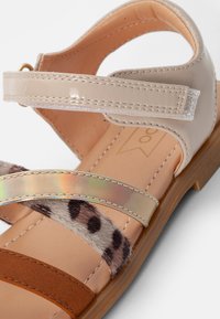 Sandal in finta pelle beige con cinturino trasparente, accento olografico e banda in camoscio marrone. Presenta un dettaglio in pelliccia con stampa animale texturizzata.