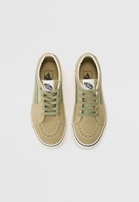 Une paire de chaussures de sport Vans beige et verte en daim avec des lacets, des semelles intermédiaires blanches et le logo "Vans Off The Wall" sur les languettes, vues de dessus.