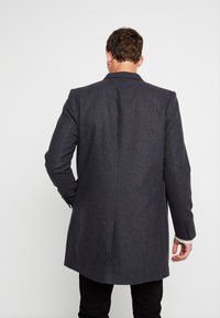 Uomo di spalle che indossa un lungo cappotto grigio scuro, pantaloni neri e una camicia chiara, in piedi contro uno sfondo chiaro e uniforme.