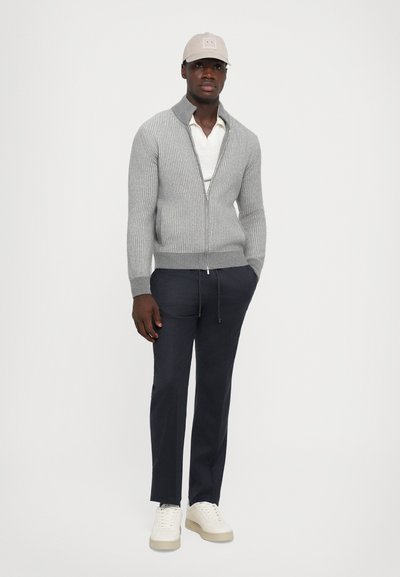 Mann mit einer hellbeigen Mütze, grauem Reißverschluss-Rippenpullover über einem weißen Poloshirt, blauen Jogginghosen mit Kordelzug und weißen Turnschuhen, steht mit entspannten Händen.
