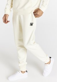 Krämfärgade bomullsjoggers med manschetter vid anklarna, svart broderad logotypdetalj på fickan, tillsammans med vita sneakers med svarta accenter.