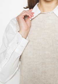 Béžový pletený svetr vrstvený přes bílou košilovou košili. Svetr má texturovaný vzor, zatímco košile má manžety a límec na knoflíky.