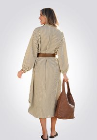 Robe rayée jusqu'aux genoux avec de longues manches ballon, taille cintrée et ceinture marron. Le mannequin tient un sac en bandoulière marron texturé, portant des sandales.