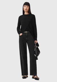 Maglione nero a maglia con un design asimmetrico, abbinato a jeans in denim neri a vita alta. Accessori includono una cintura nera e una borsa.