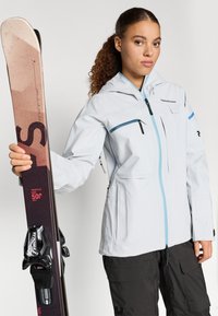 Vaaleanharmaa hiihtotakki sinisillä yksityiskohdilla, vetoketjulliset taskut ja säädettävät ominaisuudet. Musta suksi siteet kädessä vasemmalla.