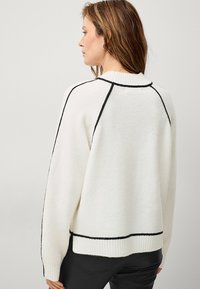Maglione bianco lavorato a maglia con dettagli neri alle cuciture e all'orlo. Presenta un colletto e polsini a coste. Taglio ampio, texture morbida.