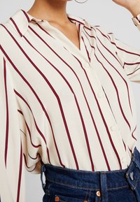 Blusa de manga corta en color crema, con rayas verticales rojas, cuello puntiagudo y cierre de botones en la parte delantera. Tejido suave y liso.