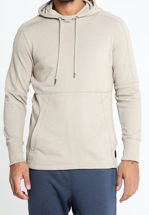 Muž v béžové mikině s kapucí, předními kapsami a šňůrkami, ve spojením s tmavě modrými kalhotami, stojící před bílým pozadím.