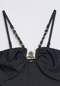 Black denim cropped top with thin adjustable straps featuring gold chain links and a central round gold clasp with a logo.