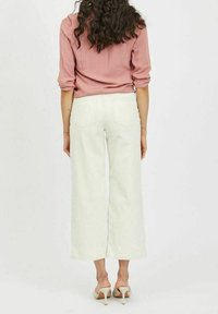 Una donna con capelli ricci indossa un top rosa a maniche lunghe, pantaloni bianchi a gamba larga e scarpe con tacco bianche, in piedi di spalle.