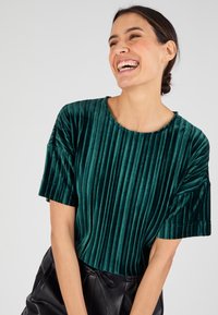 Top de terciopelo verde esmeralda con textura acanalada vertical, mangas cortas y escote redondo, combinado con pantalones de cuero negros.