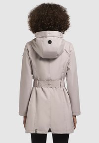 Manteau ceinturé beige clair avec capuche, présentant une taille texturée, des boutons-pression et un détail de logo noir à l'arrière. Finition en tissu lisse.