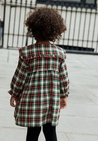 Robe à carreaux verts, rouges et blancs avec un col à volants, des manches longues et une taille froncée. Des leggings noirs complètent la tenue.