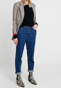 Rutigt kavaj med röda accenter, svart tröja, högmidjade blå jeans med uppvikt mudd, och ankelstövlar med ormskinnsmönster.