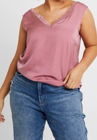 Plus-size person wearing a sleeveless dusty pink lace-trimmed V-neck top and blue high-waisted jeans, holding the top hem.