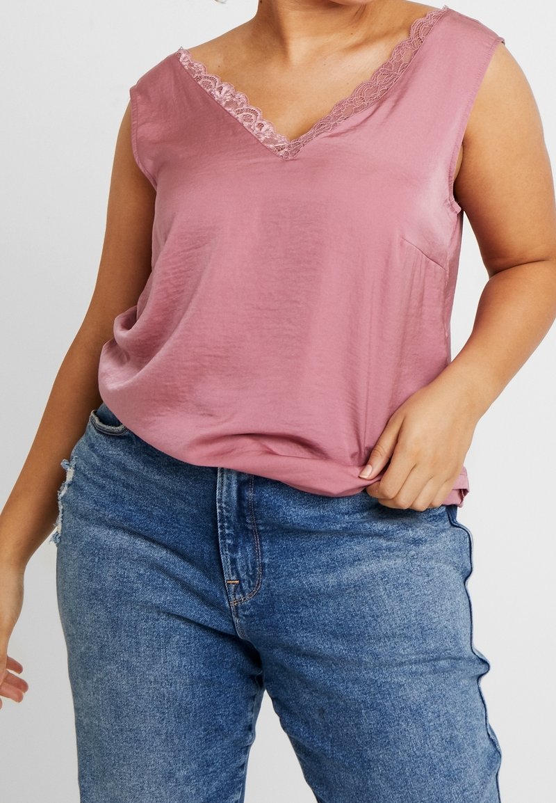 Plus-size person wearing a sleeveless dusty pink lace-trimmed V-neck top and blue high-waisted jeans, holding the top hem.