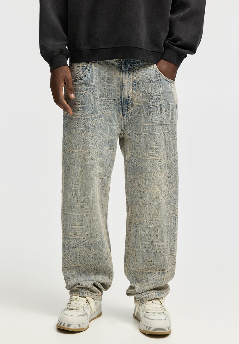 Homme portant un jean ample délavé à l'acide avec des motifs texturés, des baskets blanches et un haut noir à manches longues, les mains dans les poches.