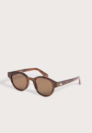 Lunettes de soleil rondes en écaille de tortue avec des verres teintés marron et des fines branches, reposant en angle sur une surface blanche.