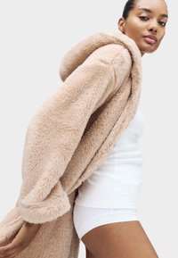 Donna in canottiera bianca e pantaloni corti che indossa un cappotto a cappuccio beige in pelo, vista di lato su uno sfondo semplice.
