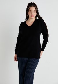 Sweater preto com decote em V e mangas longas, feito de material macio. Combinado com calças azul-marinho com riscas verticais e um ajuste relaxado.