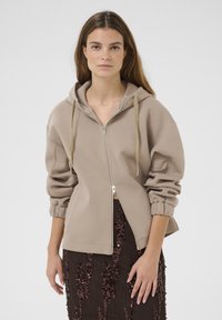 Beige vetoketjullinen huppari, jossa on vuorattu huppu, joustavat hihansuut ja rento leikkaus. Yllä tumman paljetein koristeltu hame. Sujuva, pehmeä textura.