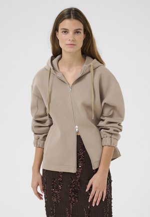 Beige Kapuzenpullover mit Reißverschluss, gefütterter Kapuze, elastischen Bündchen und einem lockeren Schnitt. Getragen über einem dunklen, glitzernden Rock. Glatte, weiche Textur.