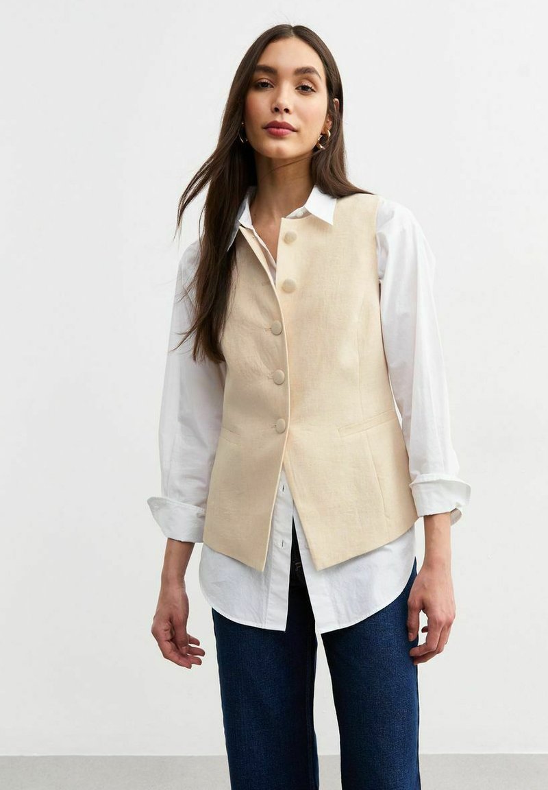 Chaleco de lino beige con un diseño a medida, que presenta cinco botones, un leve escote en forma de V y bolsillos laterales, superpuesto a una camisa blanca.