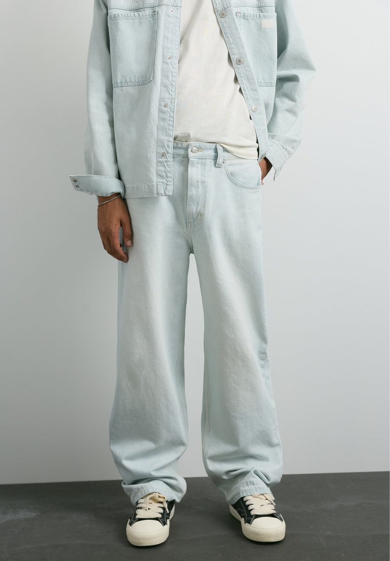 Personne portant une veste en jean bleu clair ample et un jean avec une chemise blanche et des baskets noires et blanches, debout devant un fond uni.