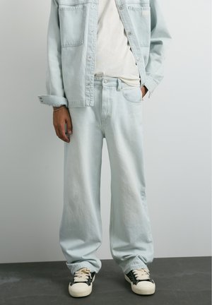 Personne portant une veste en jean bleu clair ample et un jean avec une chemise blanche et des baskets noires et blanches, debout devant un fond uni.