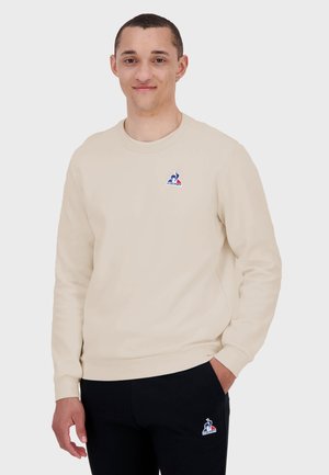 Beige Rundhals-Sweatshirt mit langen Ärmeln, auf der Brust mit einem kleinen blauen und roten Logo versehen. Weicher Stoff mit glatter Textur.
