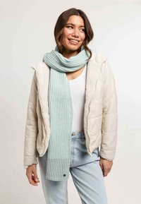 Jeune femme souriante, portant une doudoune beige claire, un haut blanc, un jean bleu clair et une épaisse écharpe tricotée bleu pâle.