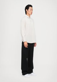 Langermet, lys beige skjorte med krage, kombinert med løse svarte pinstripe bukser og hvite sneakers.
